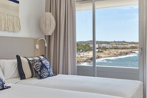 A bedroom with a large bed, a wall-mounted lamp, and a view of the beach and ocean through a large window.