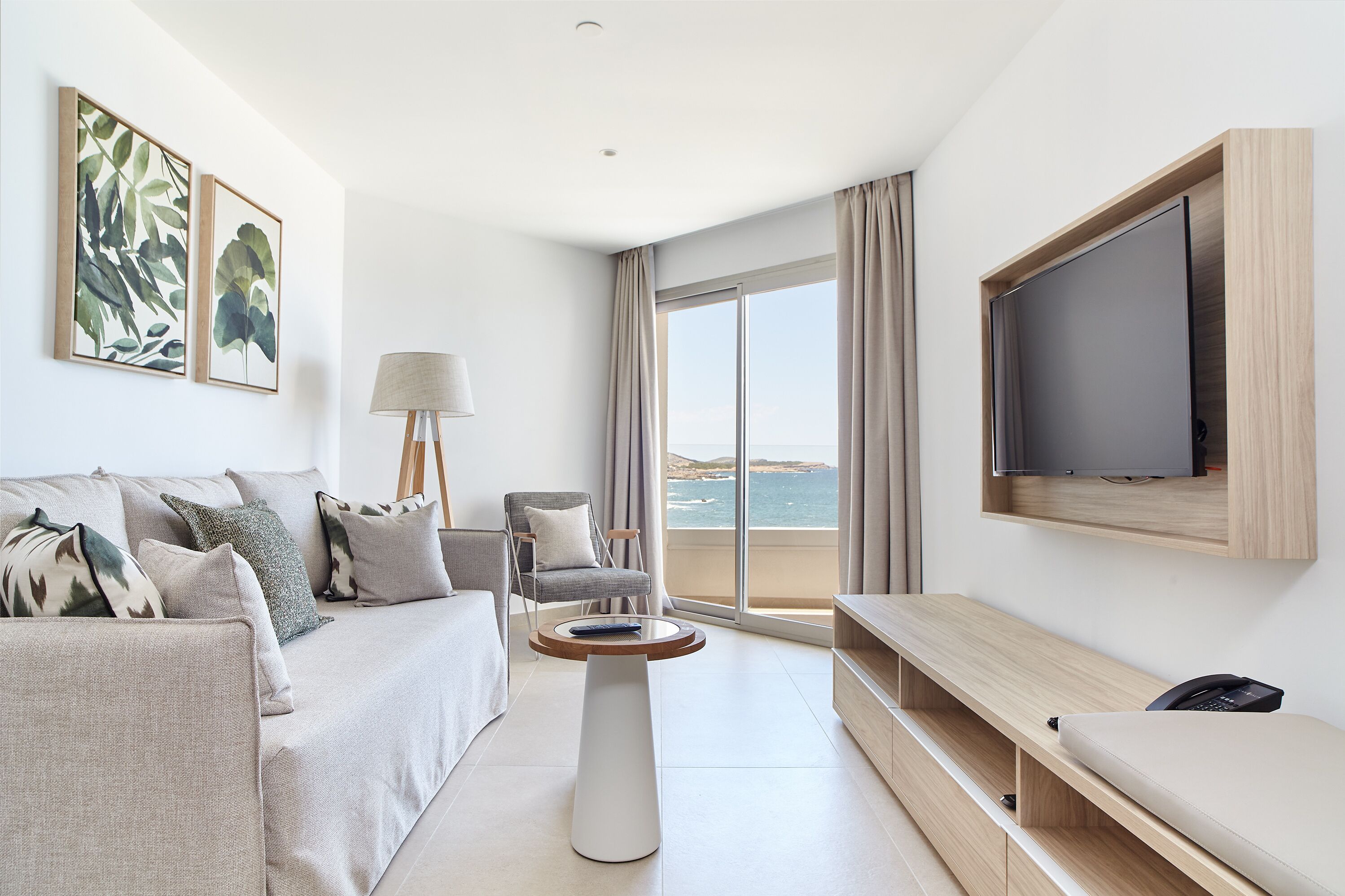 Une chambre d’hôtel moderne, dotée d’un canapé, d’une table basse, d’un téléviseur fixé au mur et offrant une vue sur la mer grâce à de grandes fenêtres.