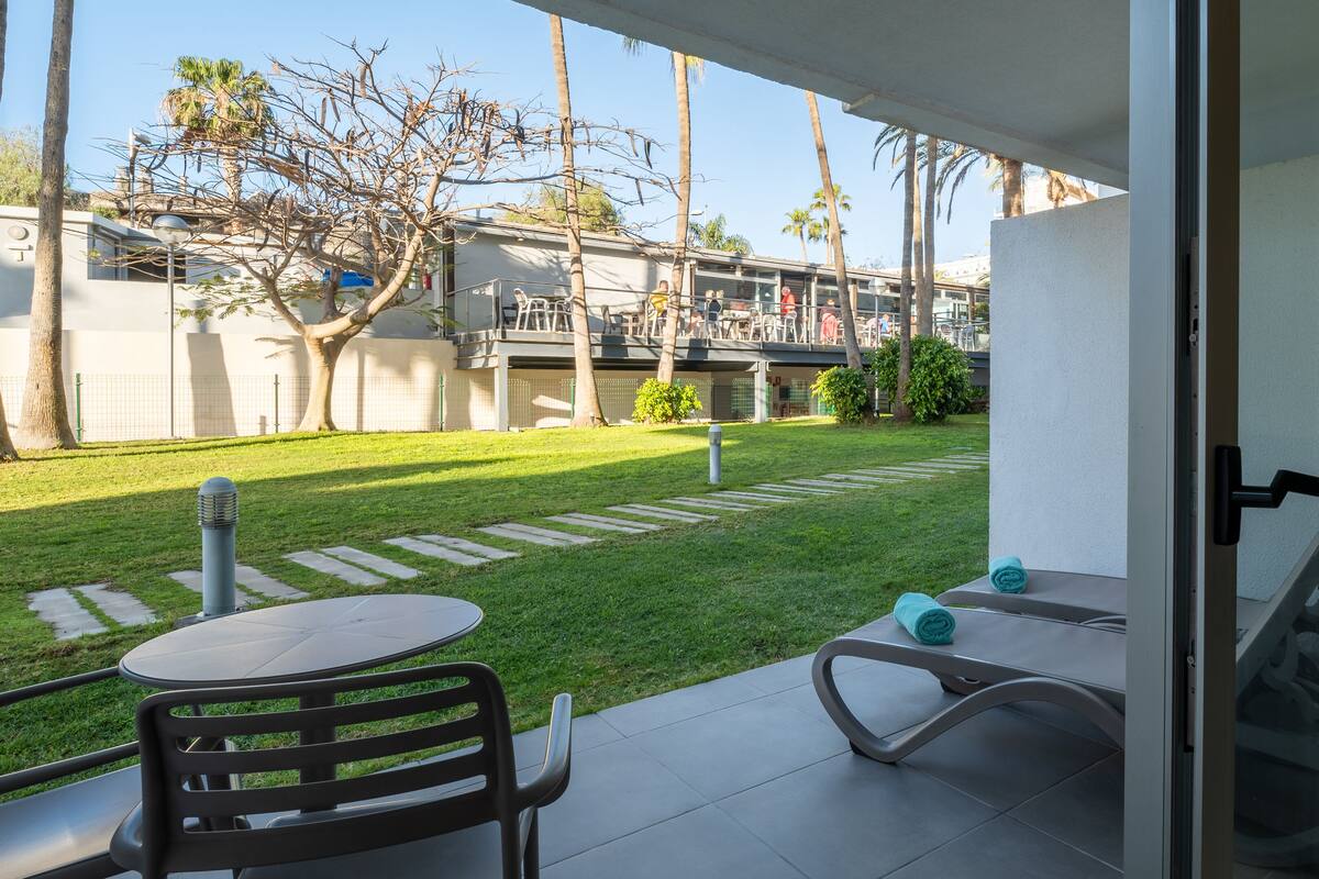 A patio with a table and chairs, a small garden, and a view of a building with people in the background.