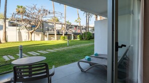 Un patio con mesa y sillas, un pequeño jardín y vistas a un edificio con gente al fondo.