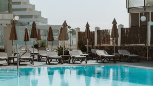 A swimming pool with lounge chairs and umbrellas, surrounded by a stone wall and modern buildings. - Apartamentos El Palmar (San Bartolomé de Tirajana)