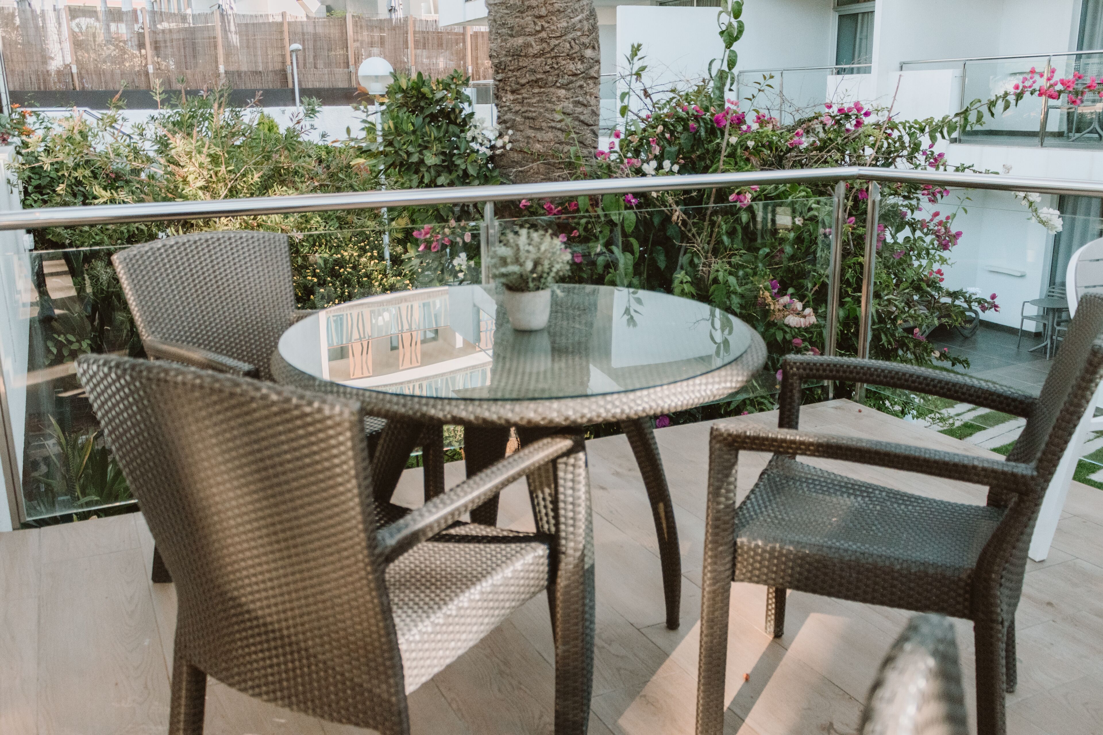 A round glass table with wicker chairs on a balcony with a metal railing.