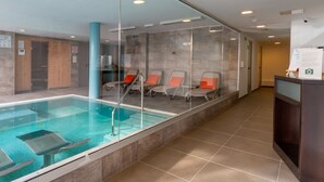 A modern indoor swimming pool with clear glass walls, a tiled floor, and several lounge chairs with orange cushions. - Apartamentos El Palmar (San Bartolomé de Tirajana)