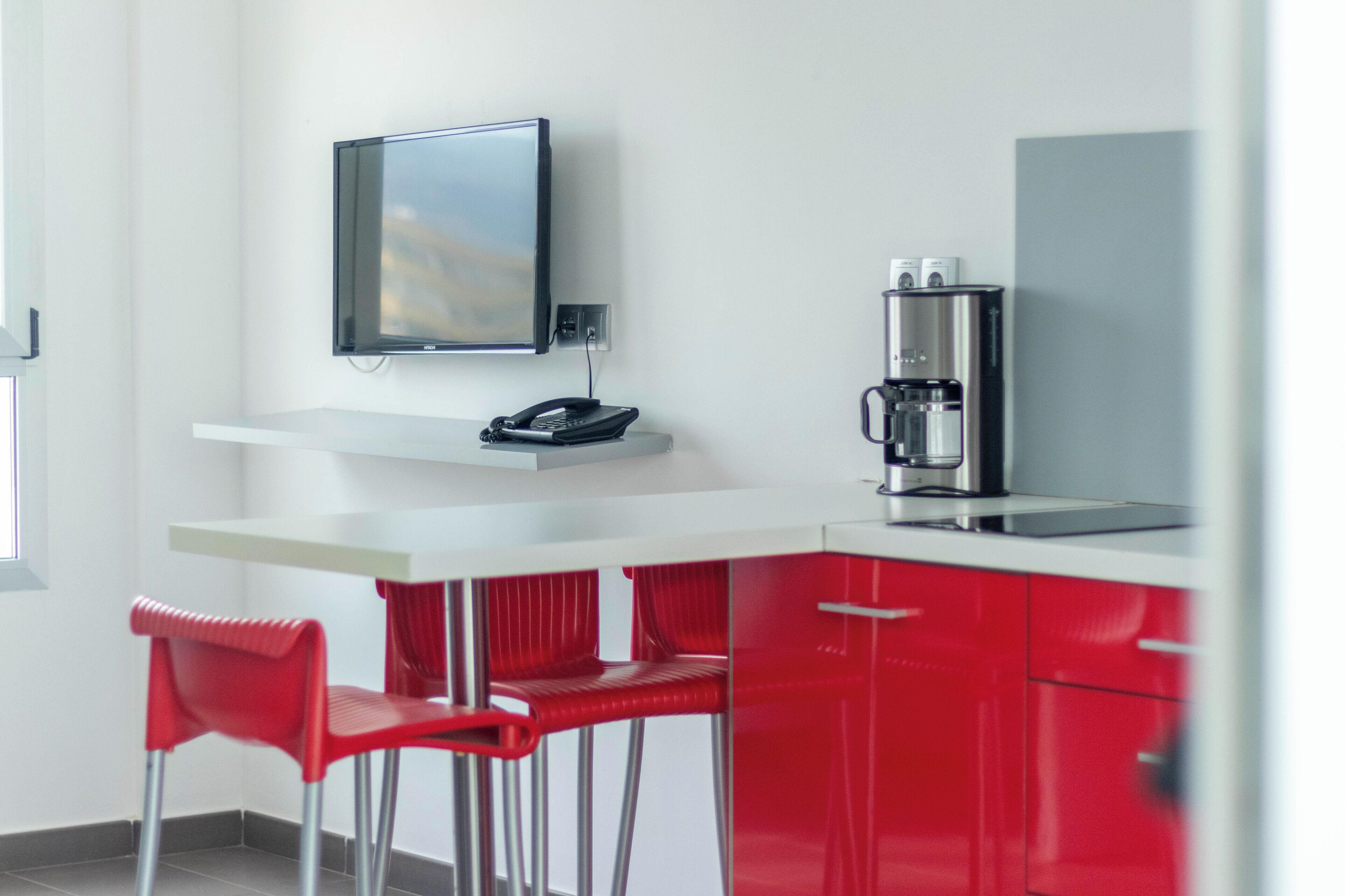 A modern kitchen with a flat-screen TV, a coffee machine, and red bar stools.