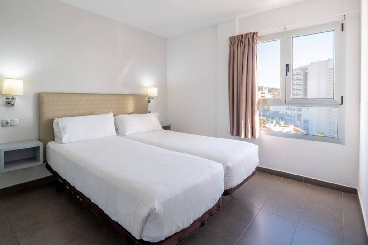 A hotel room with two beds, a headboard, bedside lamps, a small shelf, and a window with curtains.