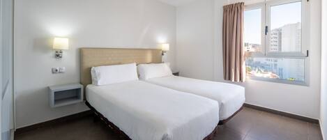 A hotel room with two beds, a headboard, bedside lamps, a small shelf, and a window with curtains.