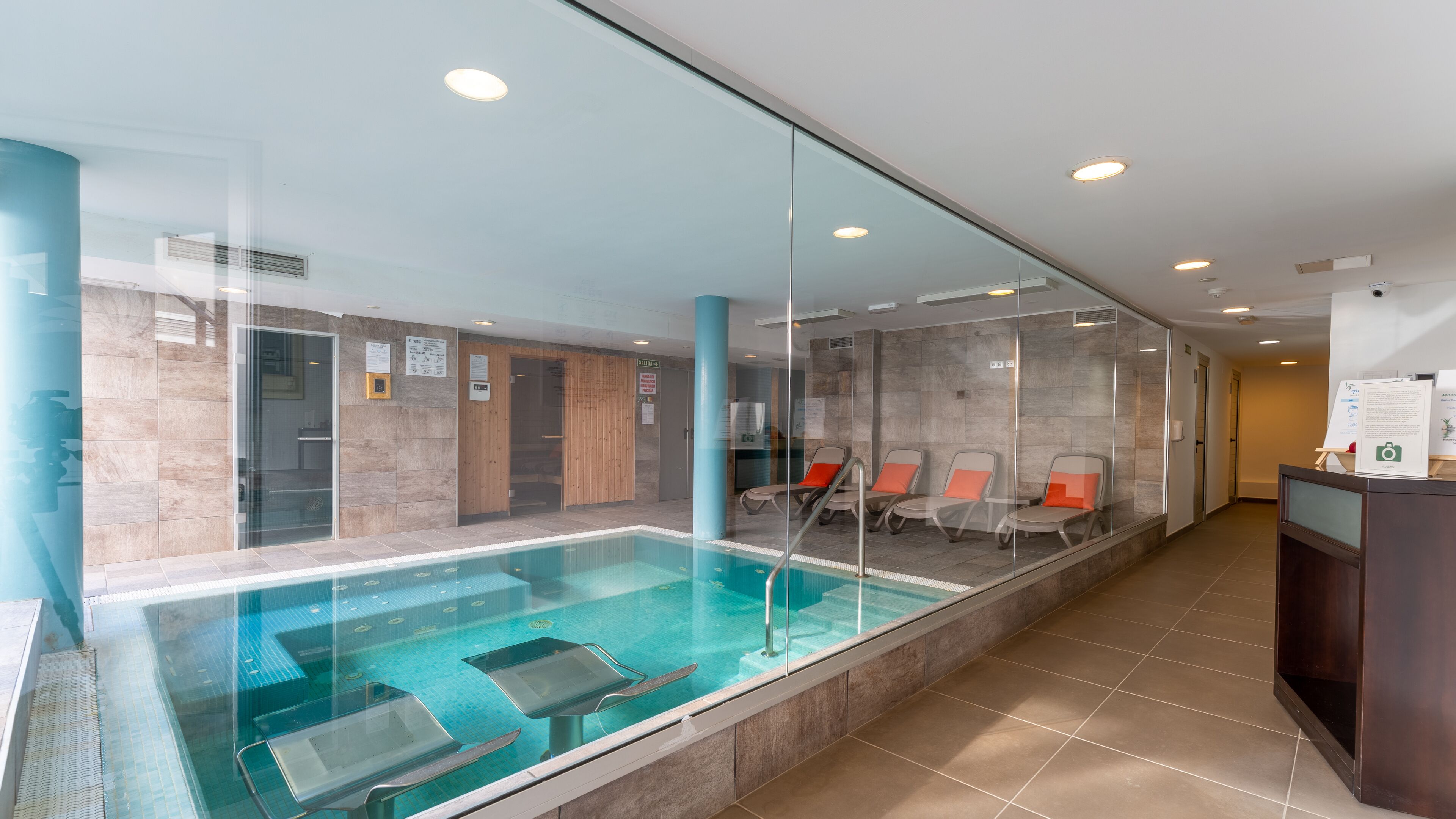 A modern indoor swimming pool with clear glass walls, lounge chairs, and a reception desk.
