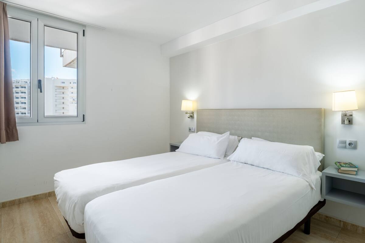 A hotel room with two single beds, a nightstand with a lamp, a small table with books, and a window with curtains.