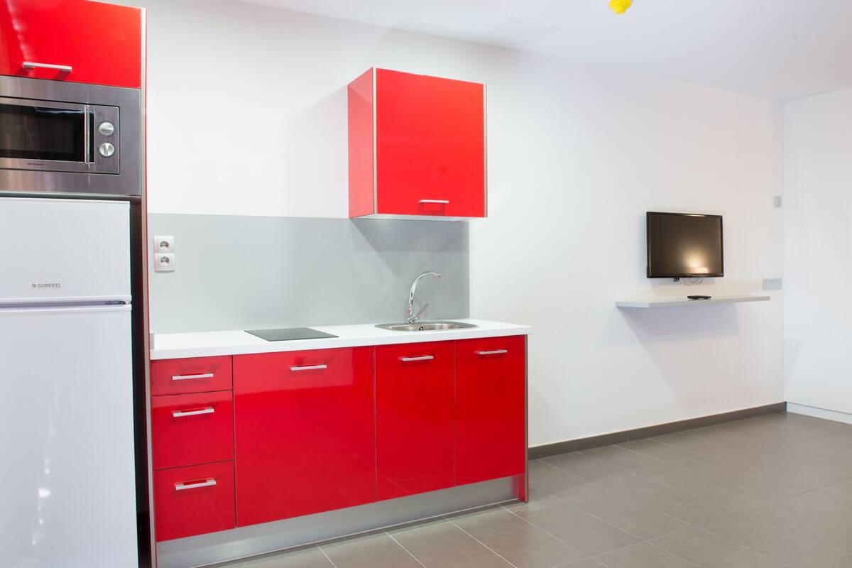 A modern kitchen with red cabinetry, a stainless steel microwave, a sink, and a flat-screen TV mounted on the wall.