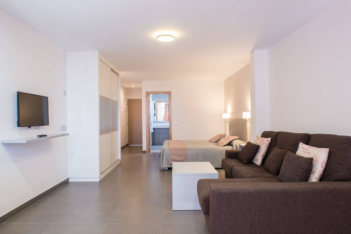 A modern living room with a sofa, a TV mounted on the wall, and a hallway leading to a bedroom.
