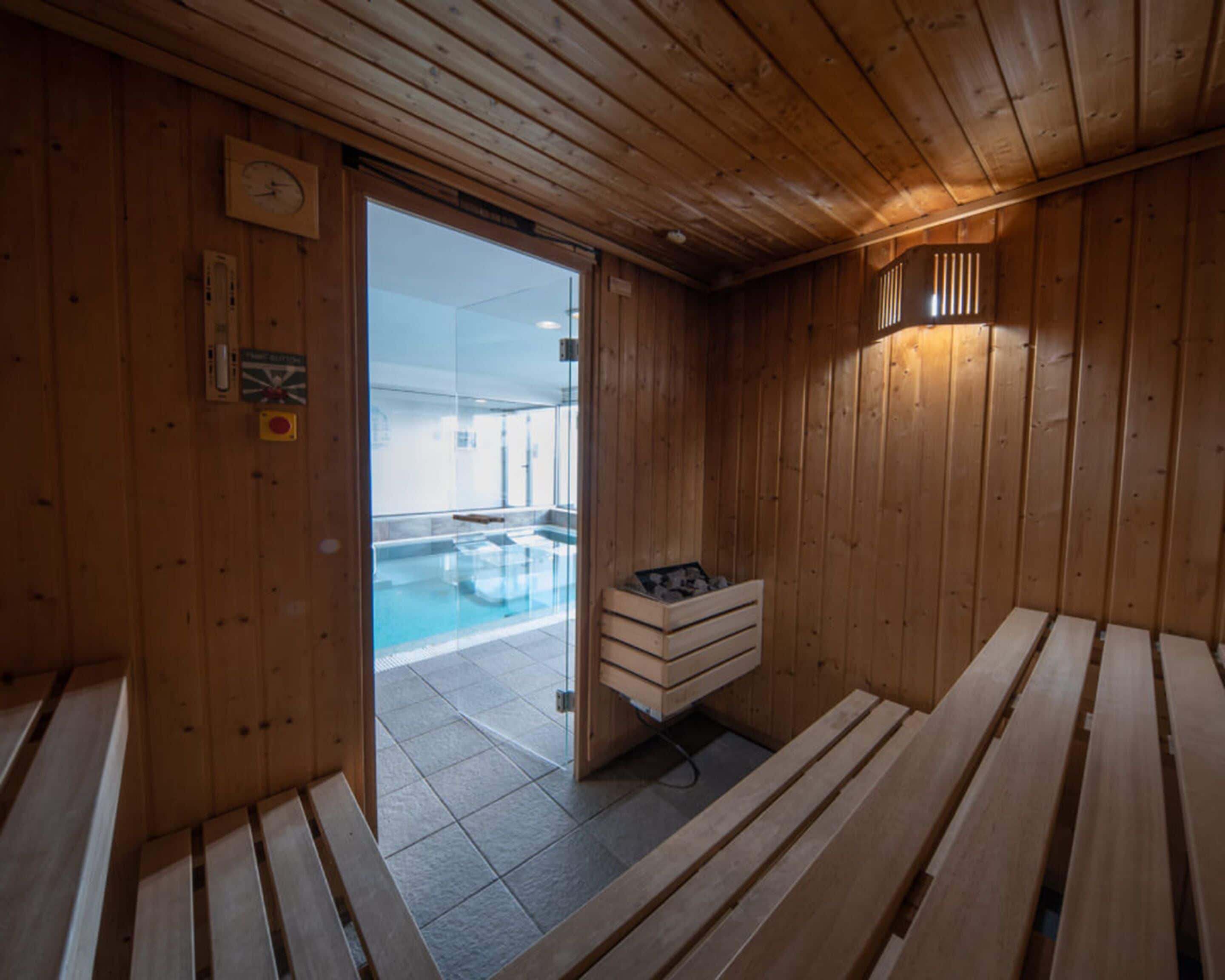 Ein Saunaraum mit Holzwänden, Bänken, einer Bank mit Heizung und einer Glastür zum Außenpoolbereich.