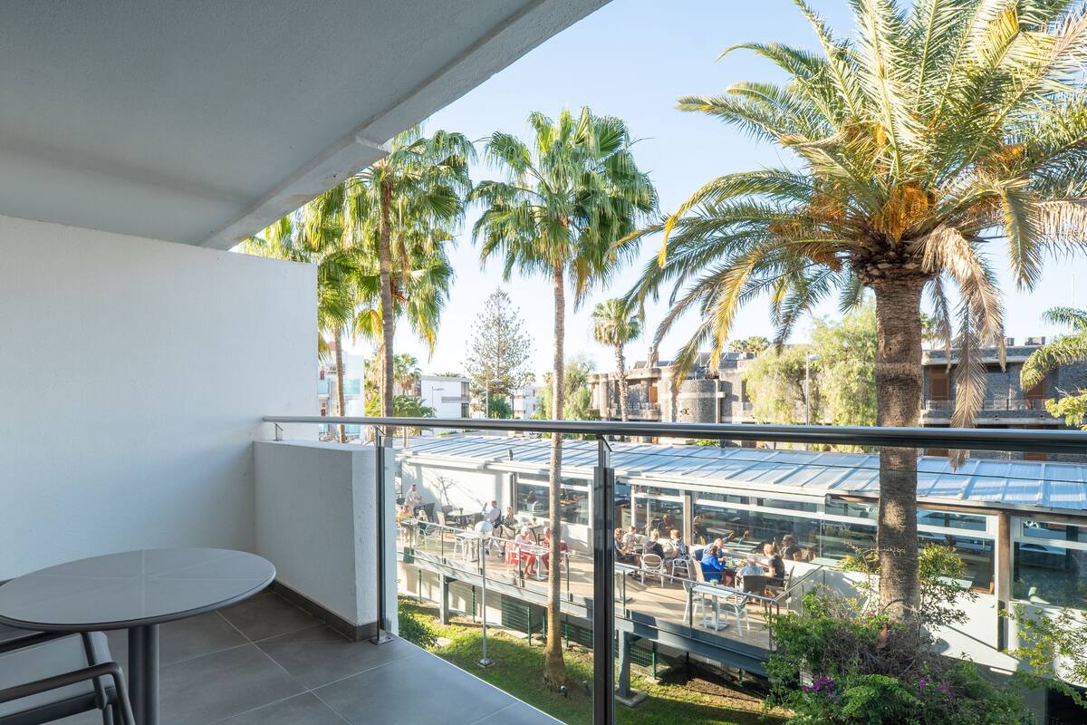 A balcony with a table and chairs, overlooking a dining area with people and palm trees.