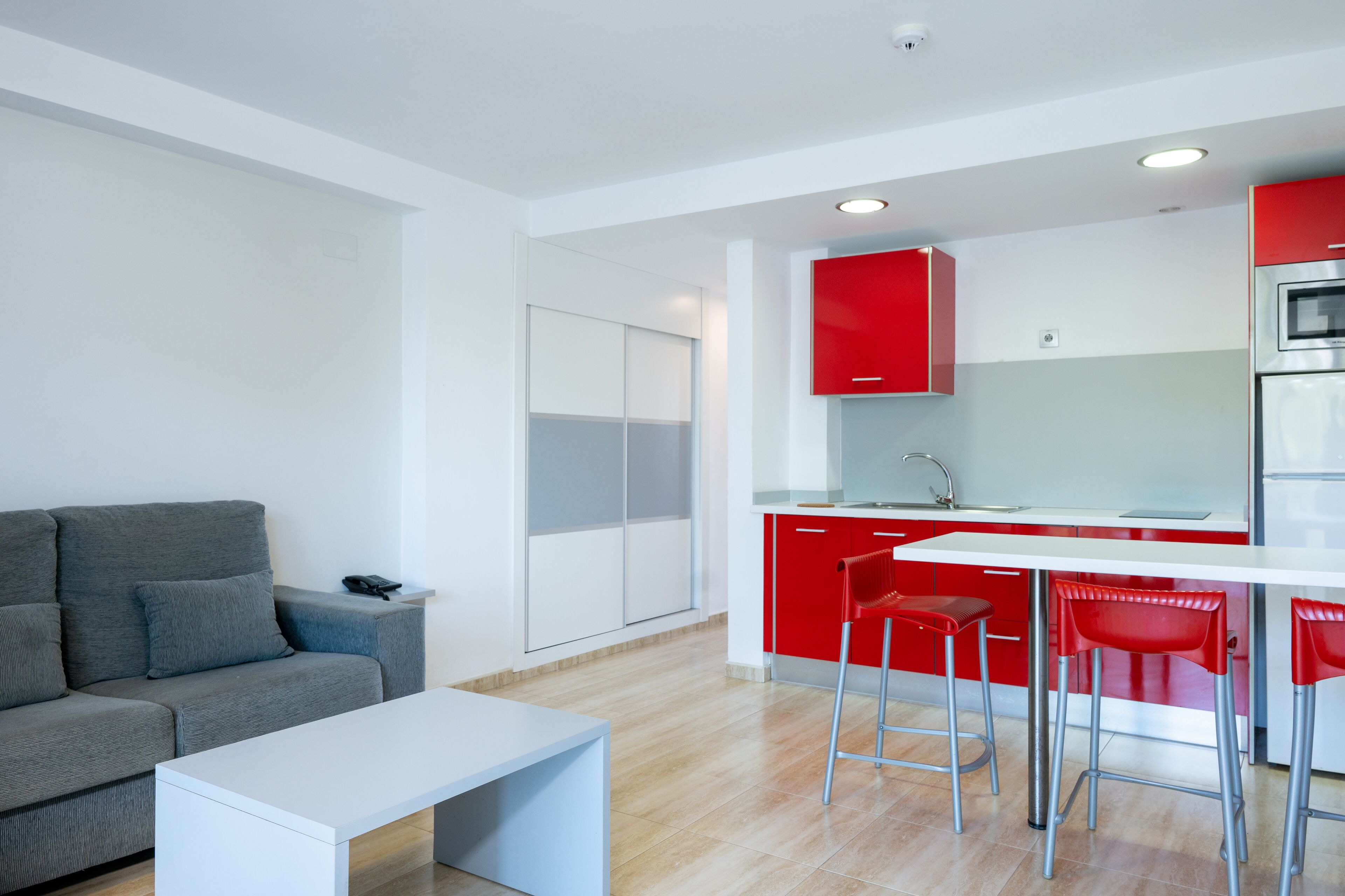 A modern kitchen with red cabinets, a white countertop, and stainless steel appliances. There is a grey sofa and a white table in the living area.