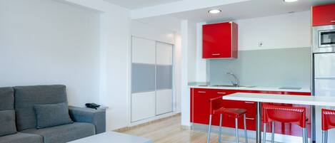A modern kitchen with red cabinets, a white countertop, and stainless steel appliances. There is a grey sofa and a white table in the living area.