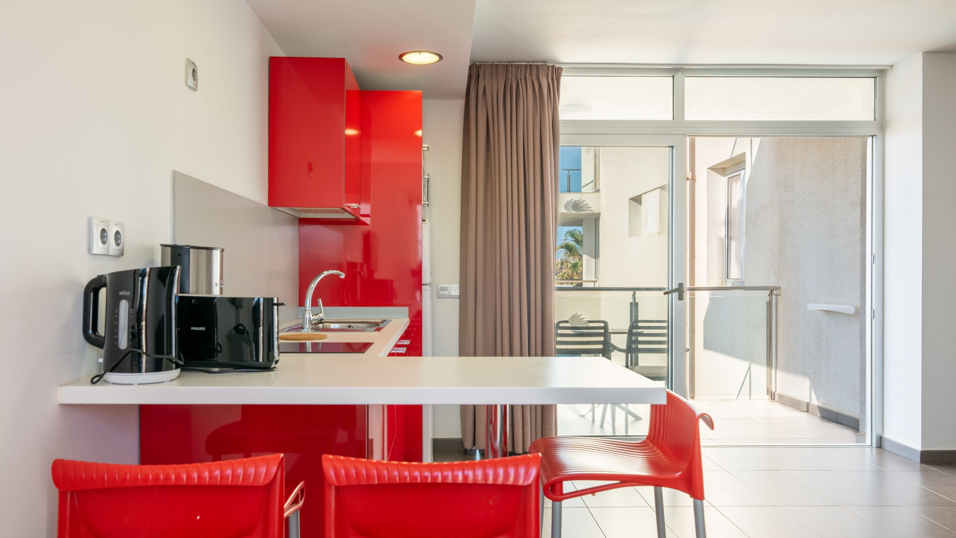 A modern kitchen with red cabinetry, a black coffee maker, and a dining area with red chairs.