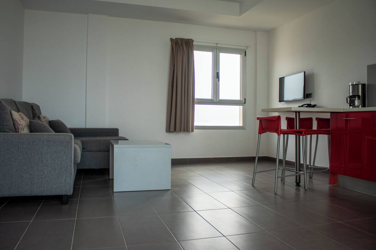 A modern living room with a grey sofa, a small white side table, and a red bar area with stools.