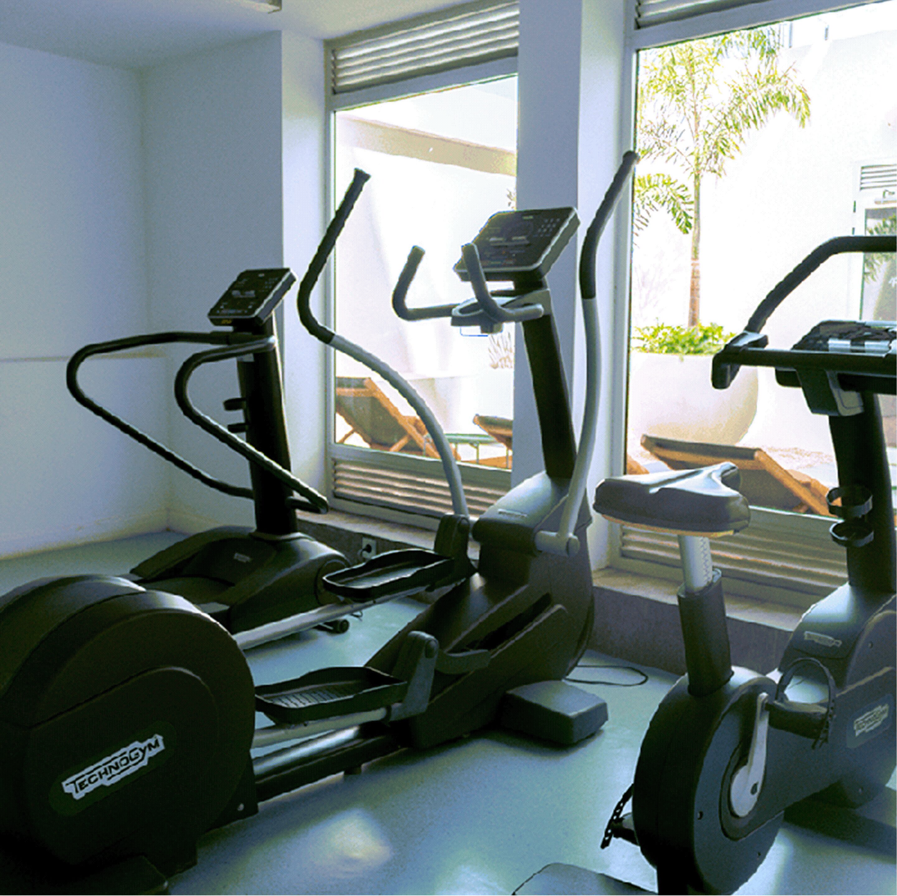 Un gimnasio con bicicletas estáticas y cintas de correr, amplias ventanas y vistas a mobiliario exterior.