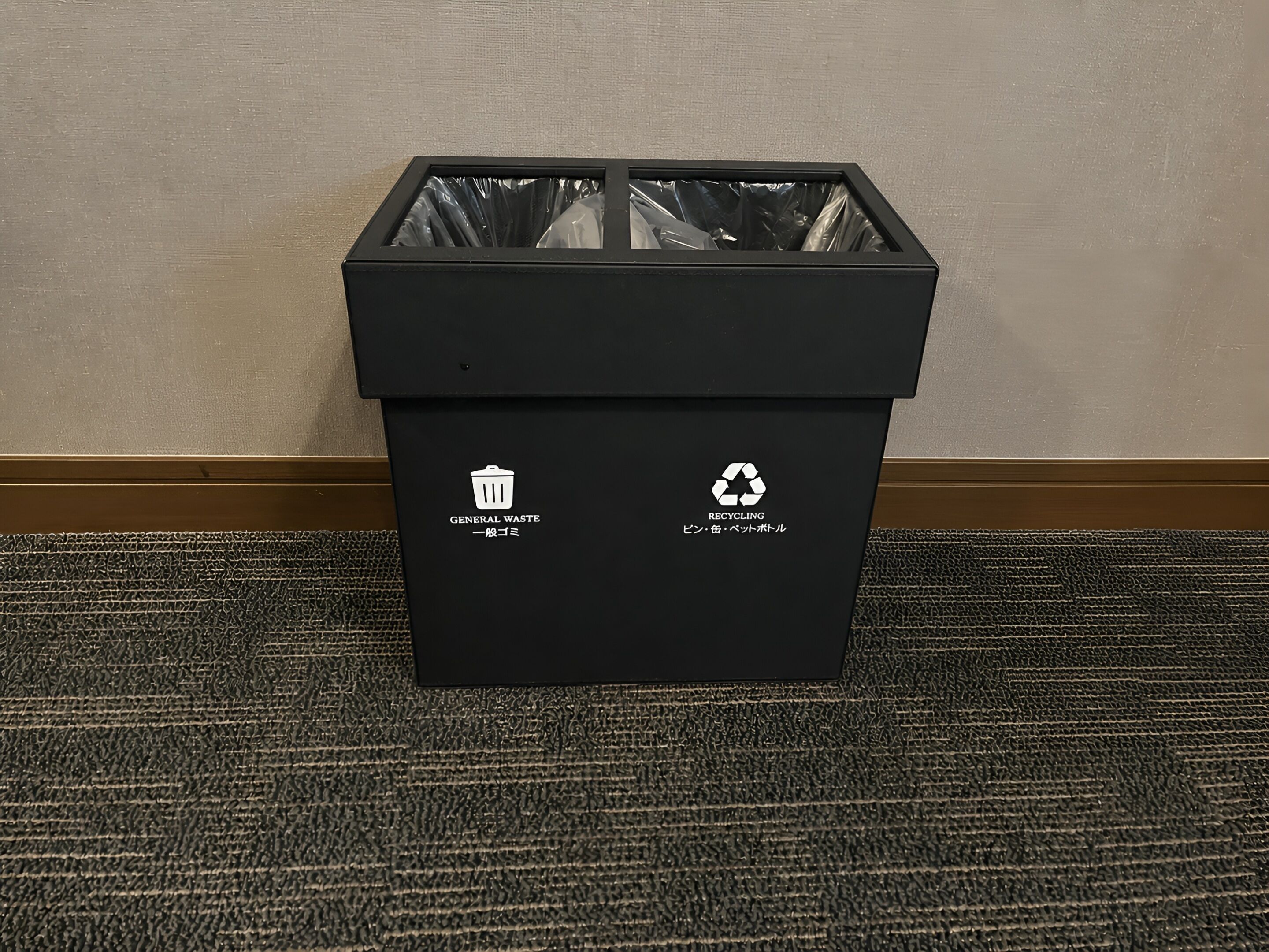 A black trash bin with recycling and general waste symbols, placed on a textured carpet against a plain wall.