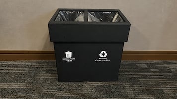 A black trash bin with recycling and general waste symbols, placed on a textured carpet against a plain wall.