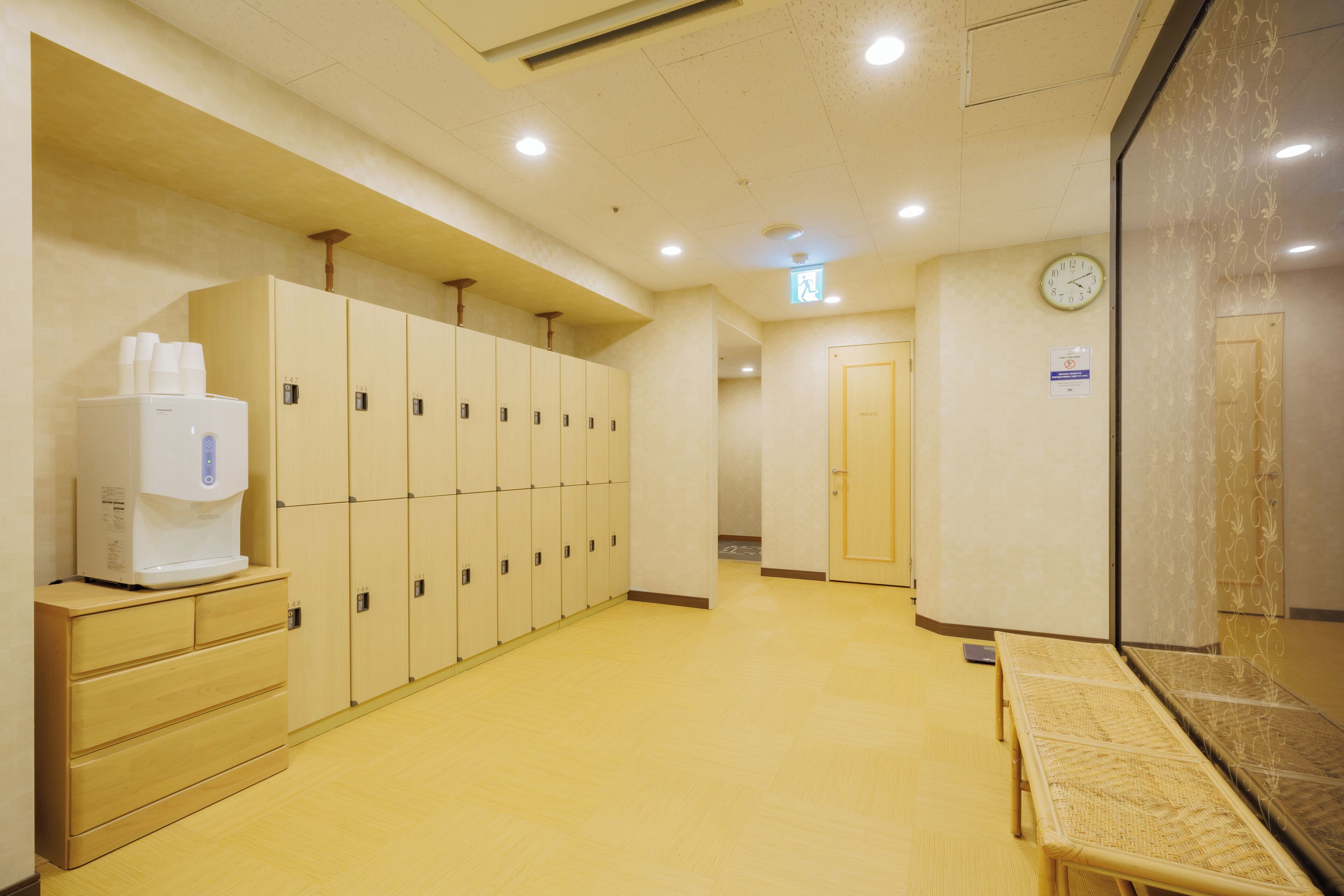 A hotel corridor with lockers, a coffee machine, a clock, and a seating area.