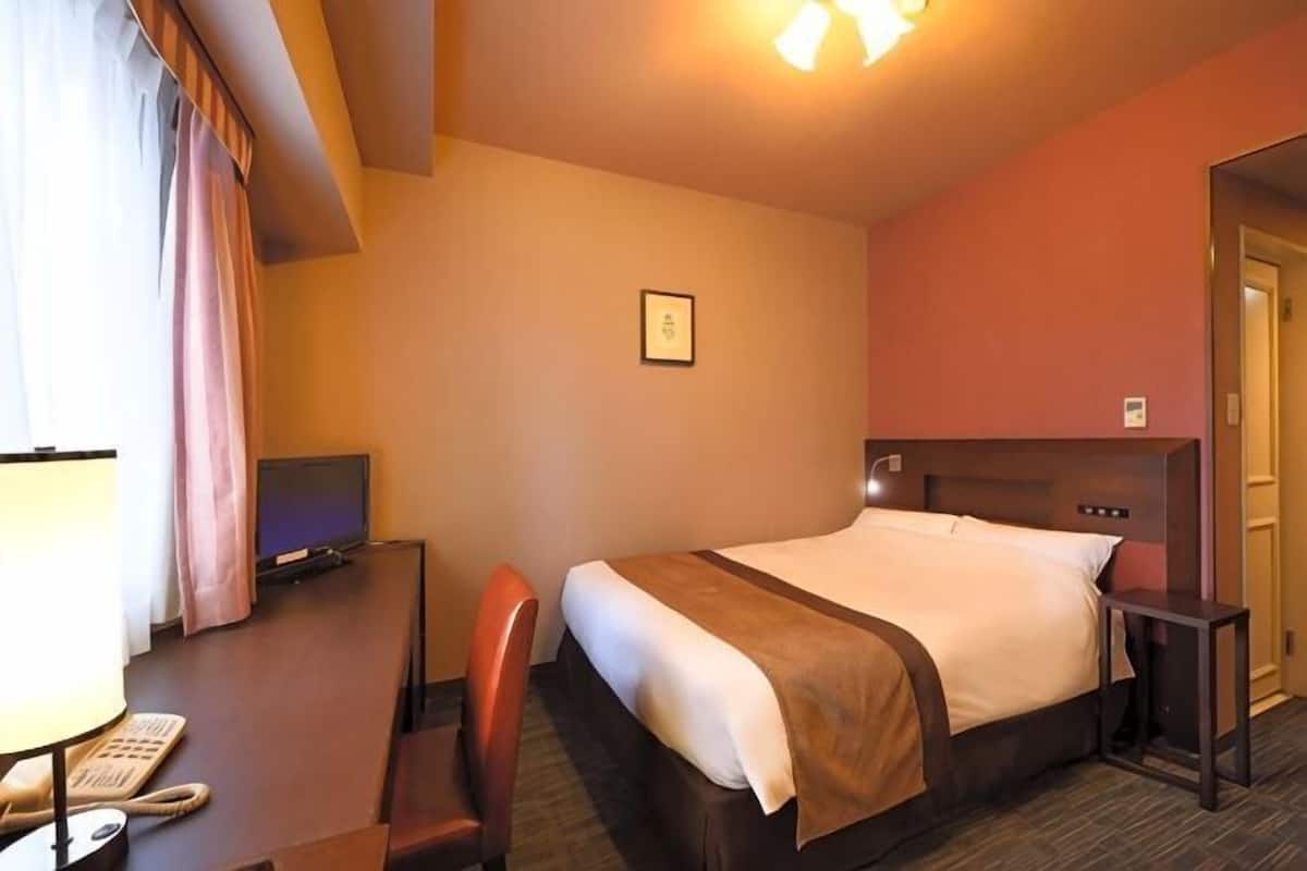 A hotel room with a bed, a desk with a computer, a chair, a lamp, and a television.