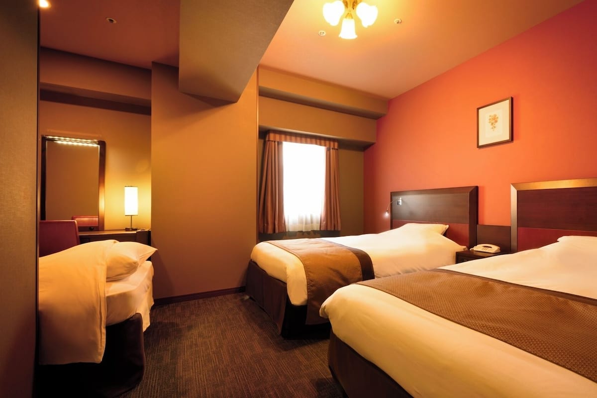 A hotel room with two beds, a bathroom, a mirror, a bedside table with a lamp, and a picture on the wall.