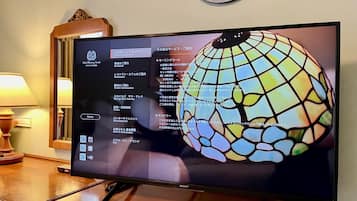 Een hotelkamer met een grote flatscreen televisie waarop een menu wordt weergegeven, een houten bijzettafeltje met een lamp en een decoratieve wandklok.