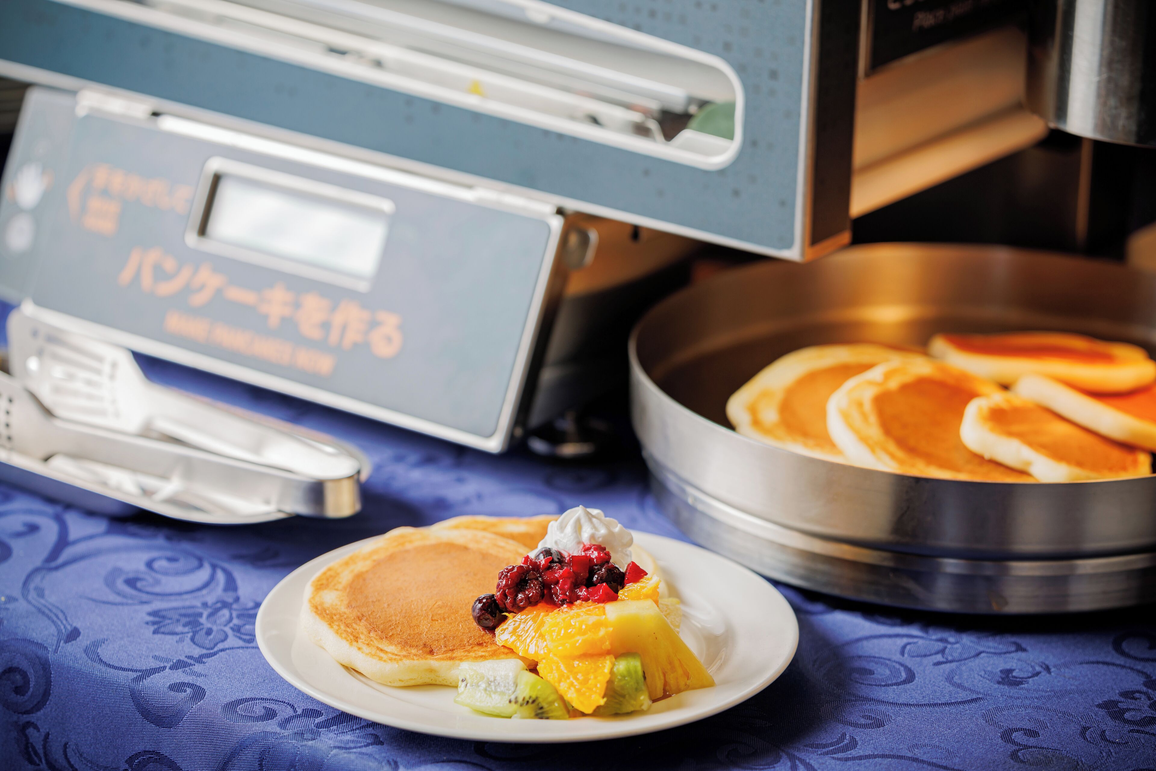Un desayuno con tortitas, fruta y crema batida en un plato, una pila de tortitas en una sartén, y una cafetera al fondo.
