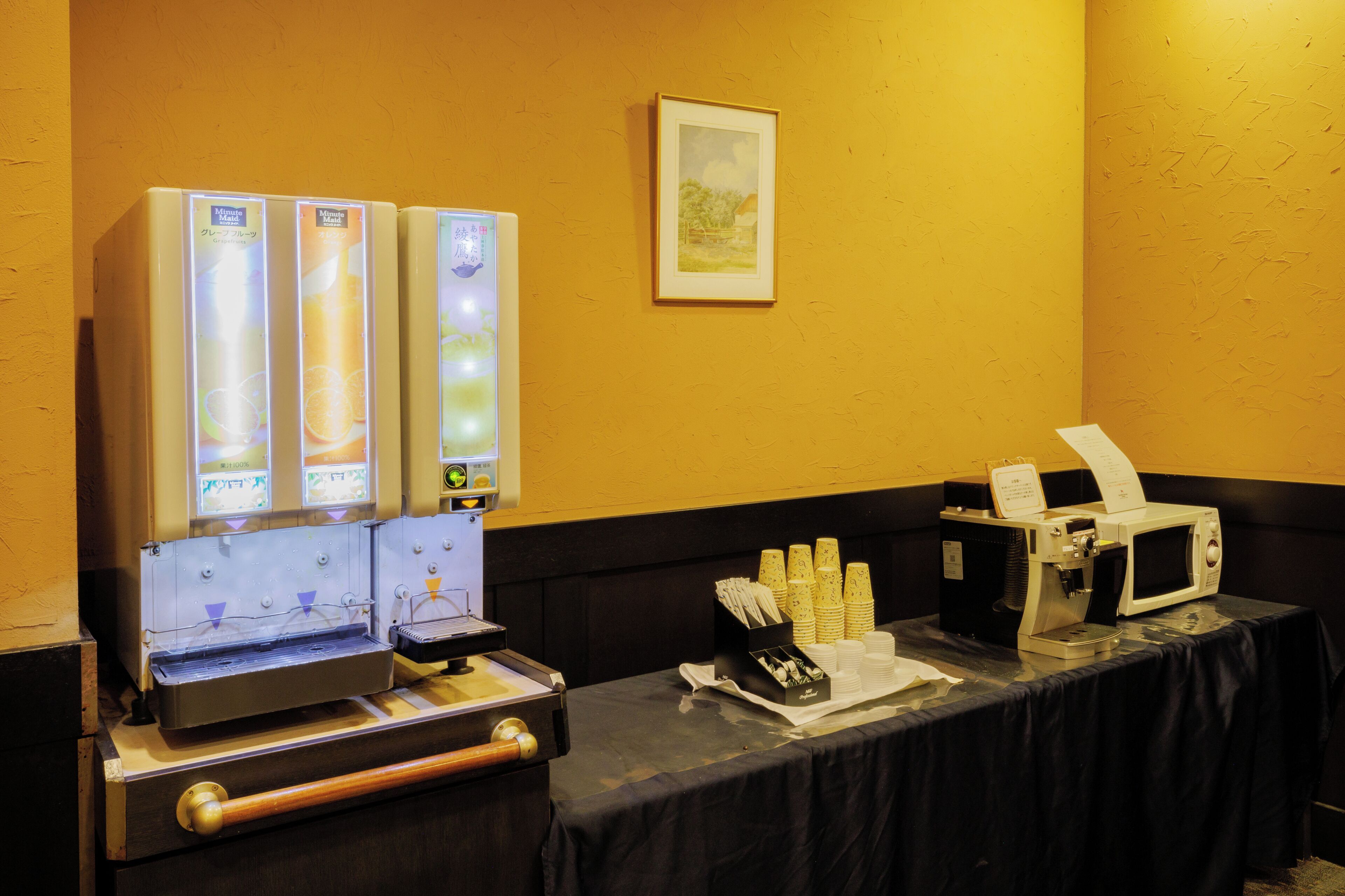 A buffet setup with beverage dispensers, a microwave, and a table with stacked plates and napkins.