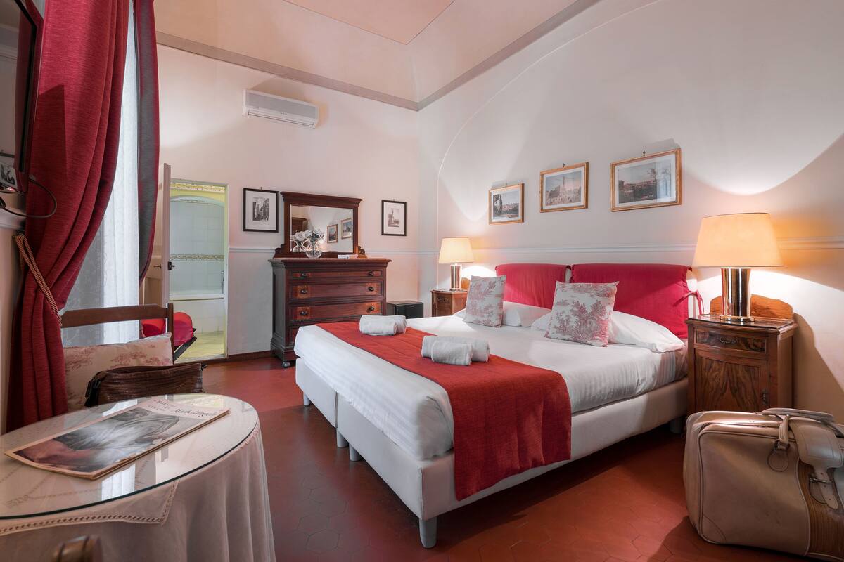 A hotel room with a large bed, a wooden dresser, a table with a newspaper, and a suitcase.