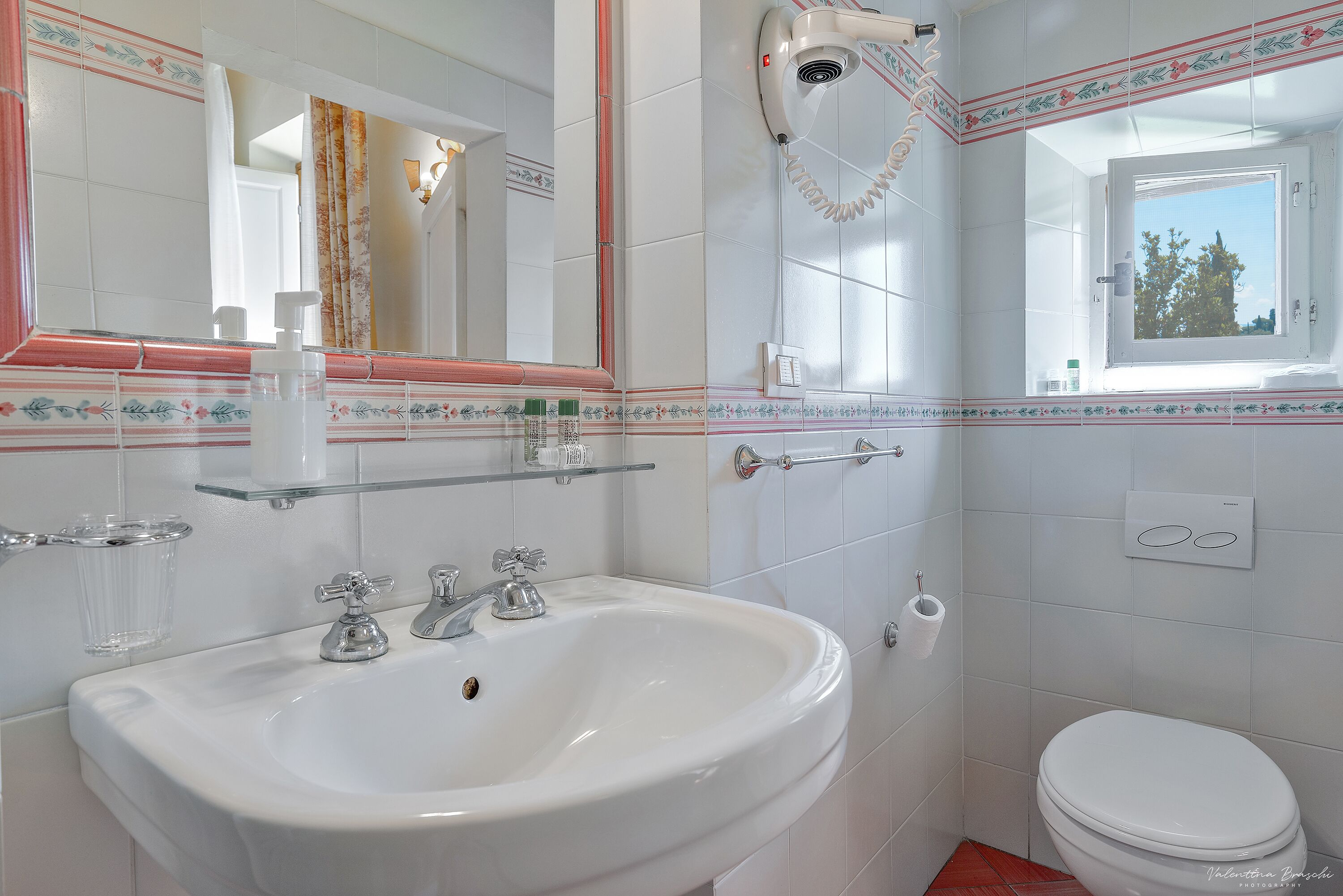 A bathroom with a white sink, two faucets, a mirror, a hairdryer, a window, and a toilet.