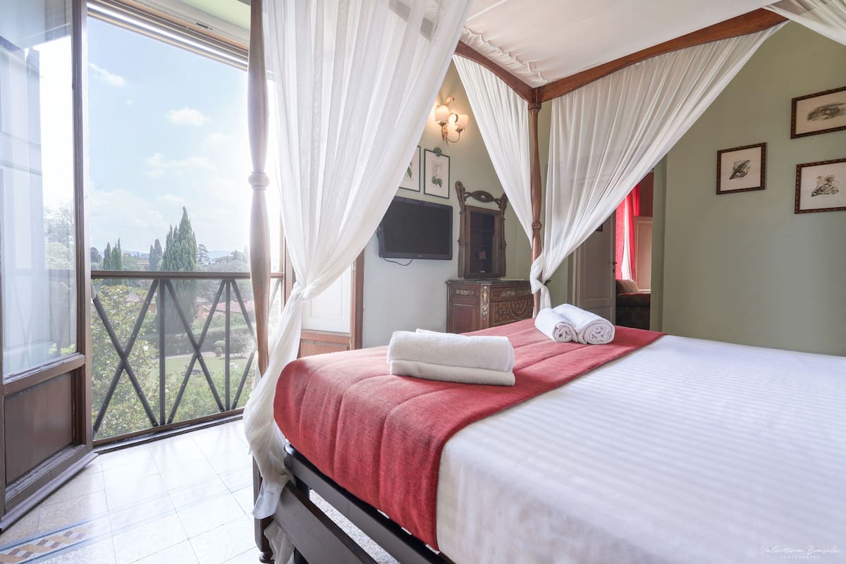 Un dormitorio con una cama, una colcha roja, cortinas blancas, un televisor y cuadros en la pared.