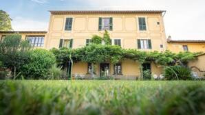 Un bâtiment à deux étages, doté d’une façade jaune et de volets verts, entouré d’une végétation luxuriante et d’un jardin bien entretenu.
