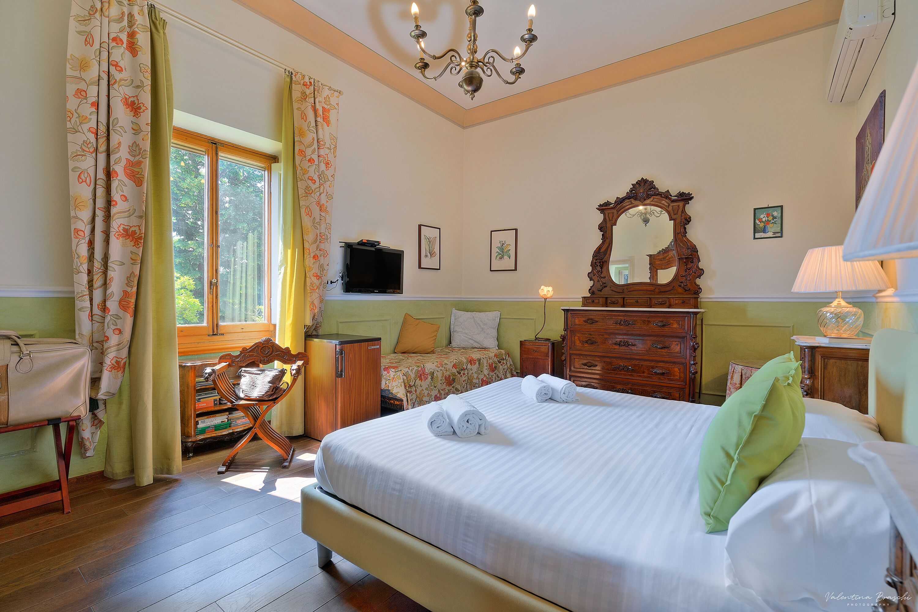 Une chambre à coucher avec un grand lit, une commode en bois, un bureau avec une chaise, une télévision et une fenêtre avec des rideaux.
