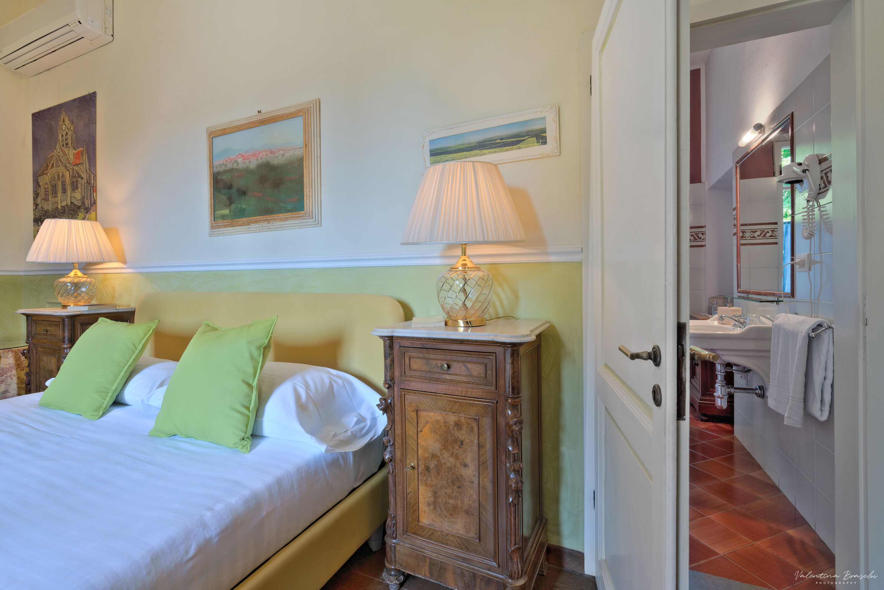 A bedroom with a bed, two bedside tables with lamps, a wooden cabinet, and a bathroom visible through an open door.