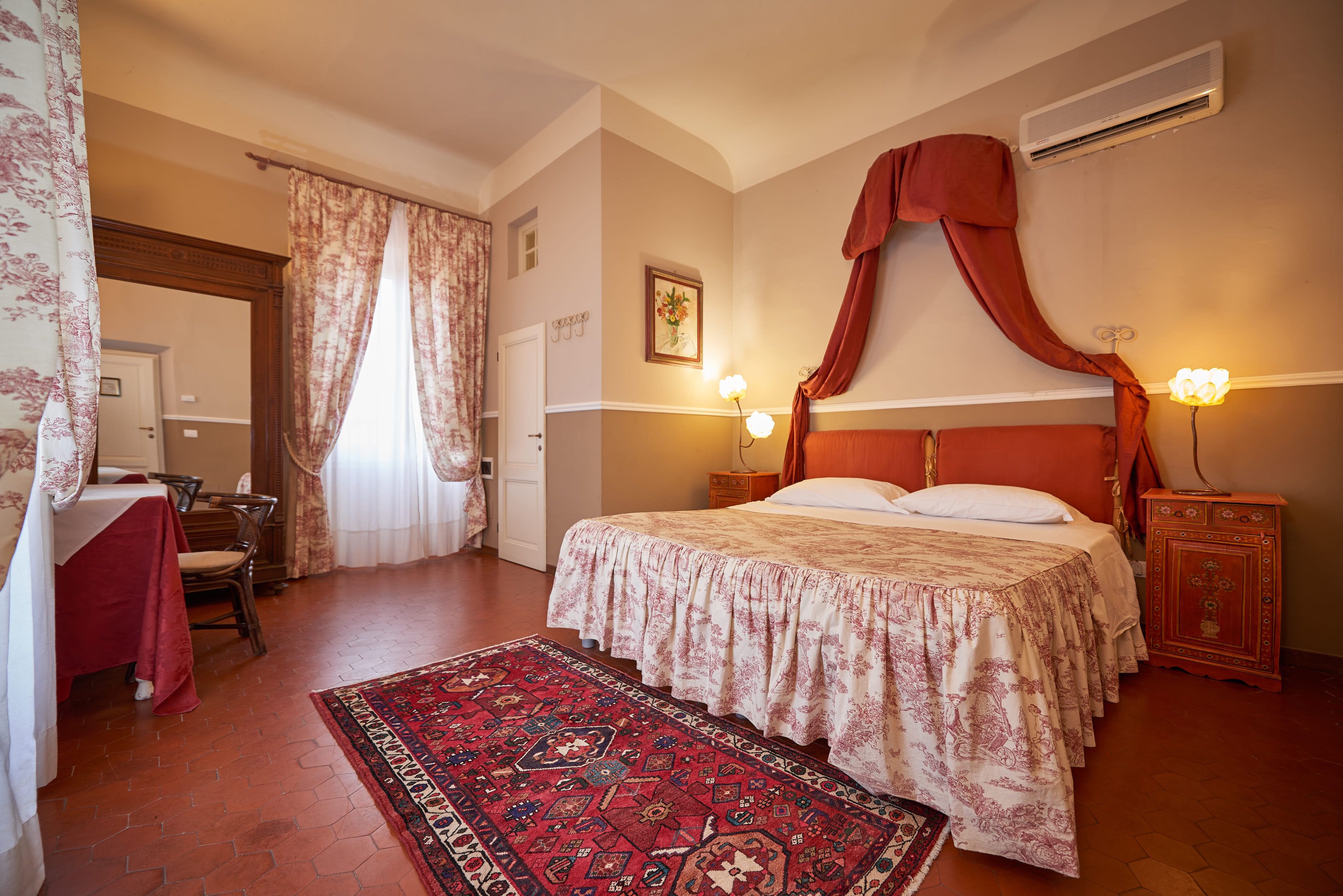 A bedroom with a large bed, a red canopy, a nightstand with a lamp, a wooden cabinet, and a patterned rug.