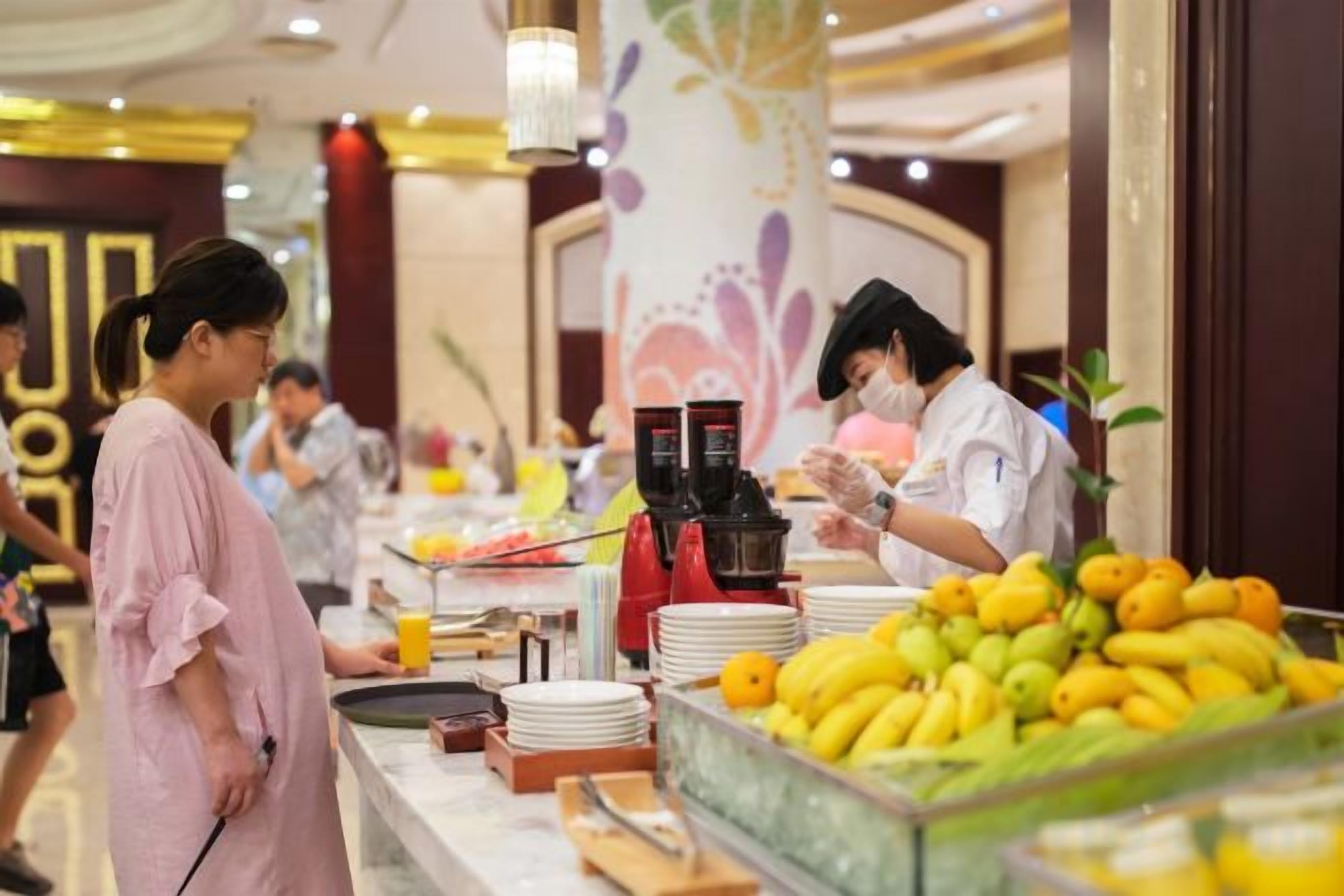 A buffet area with a variety of fruits and beverages, a counter with a staff member, and a decorative wall in the background.