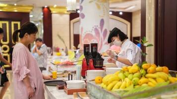 A buffet area with a variety of fruits and beverages, a counter with a staff member, and a decorative wall in the background.