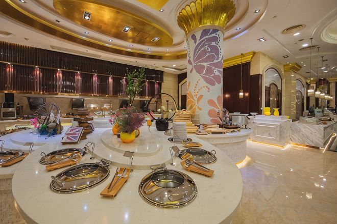 A luxurious hotel lobby with a central round table set for dining, a large decorative pillar, and a reception area with a counter and seating.