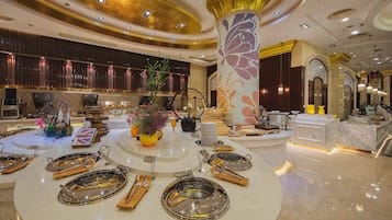 A luxurious hotel lobby with a central round table set for dining, a large decorative pillar, and a reception area with a counter and seating.