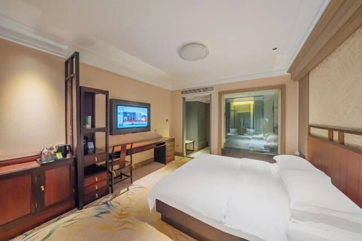A hotel room with a large bed, a desk with a computer, a television, and a glass door leading to a bathroom.