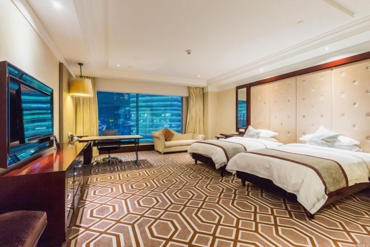 A hotel room with two beds, a TV, a desk, and a large window with a city view.