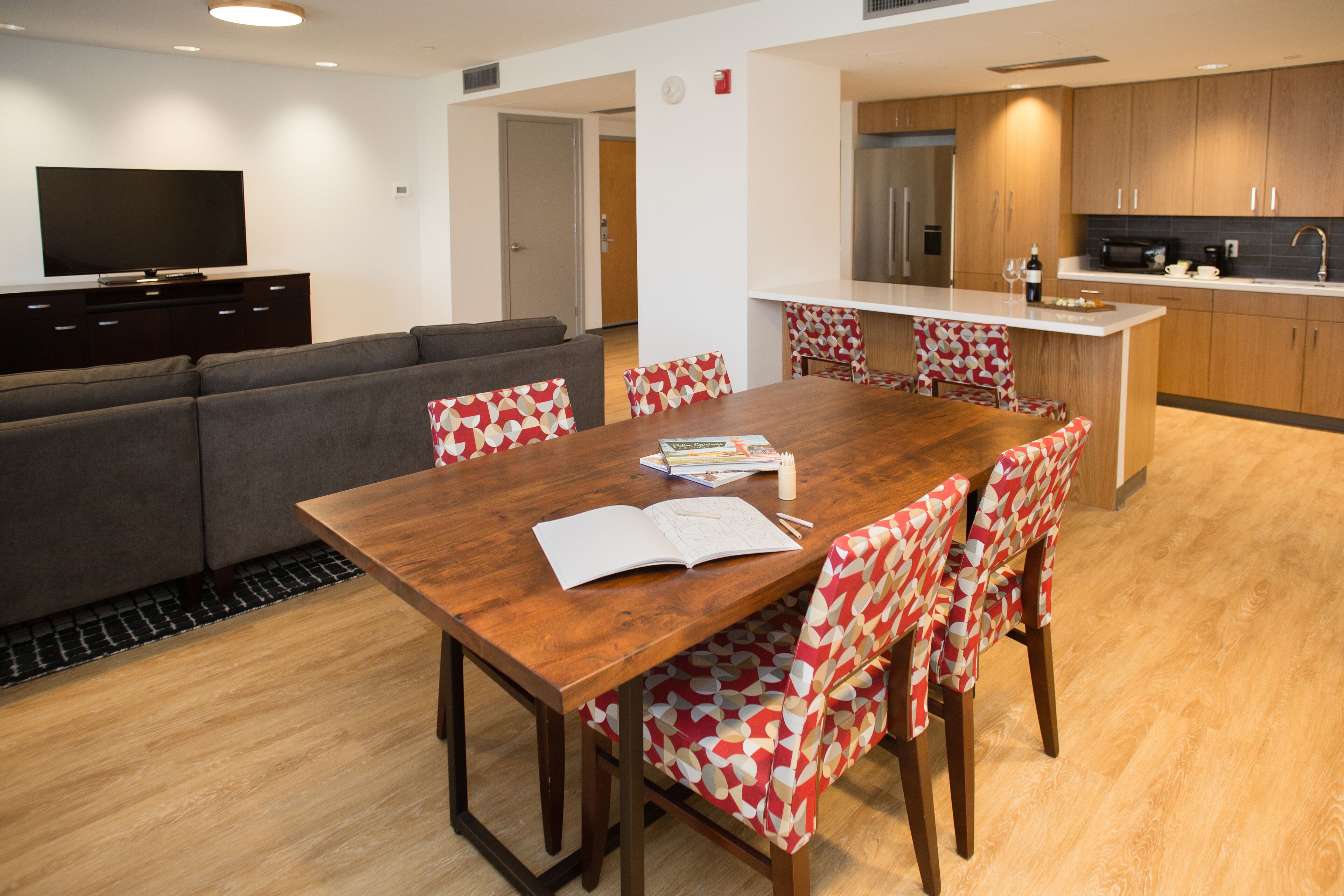 A modern living room with a wooden dining table, patterned chairs, a grey sofa, and a flat-screen TV on a dark cabinet.