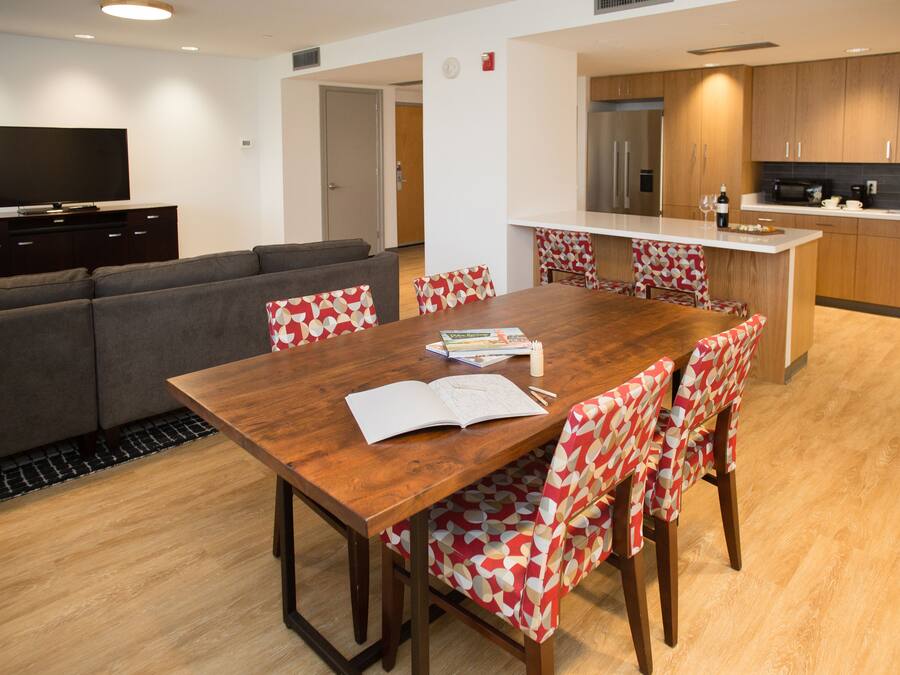 A modern living room with a wooden dining table, patterned chairs, a grey sofa, and a flat-screen TV on a dark cabinet.