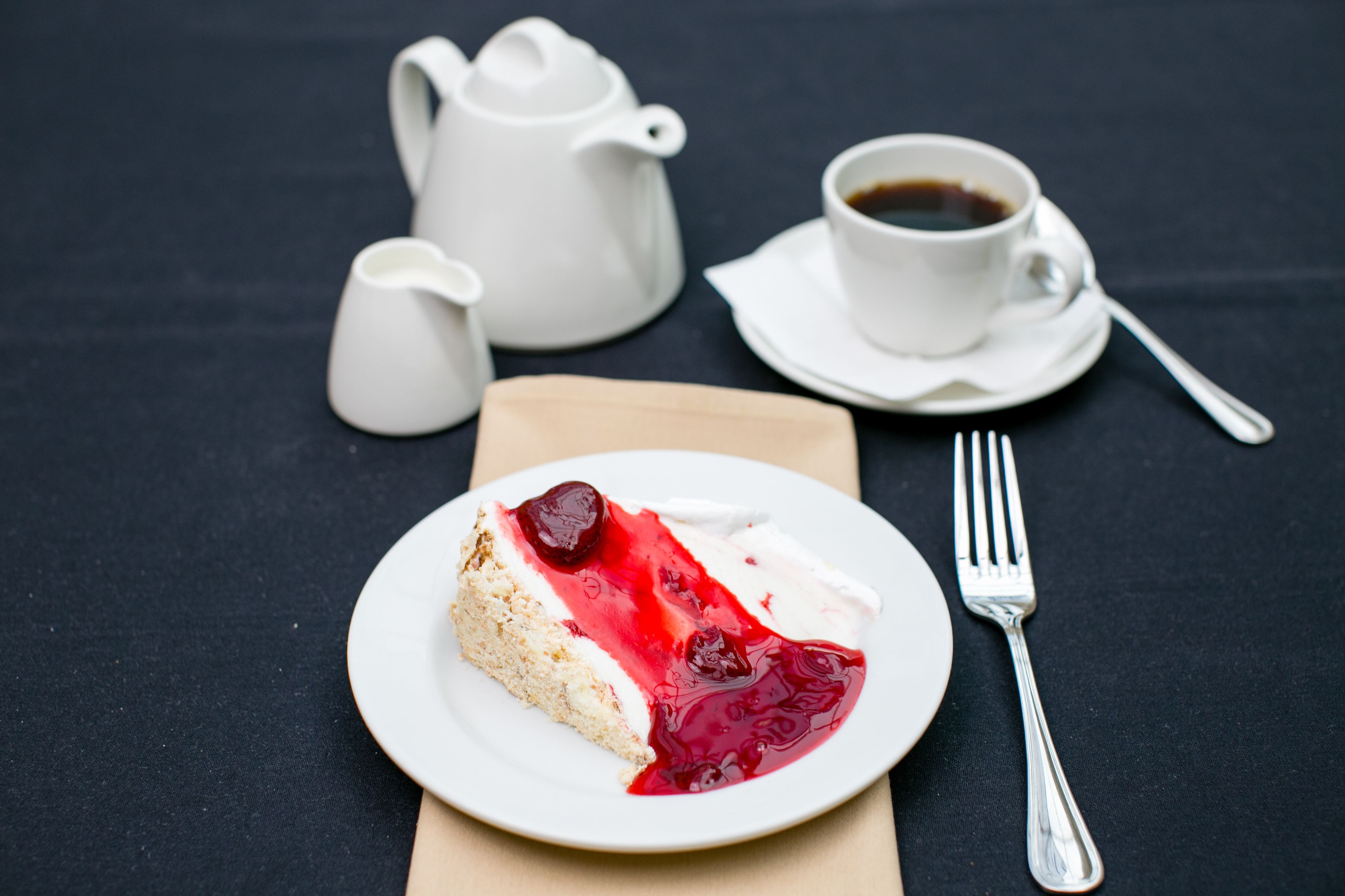A slice of cake with red fruit topping on a white plate, a white teapot, a cup of coffee, and a fork on a dark table.