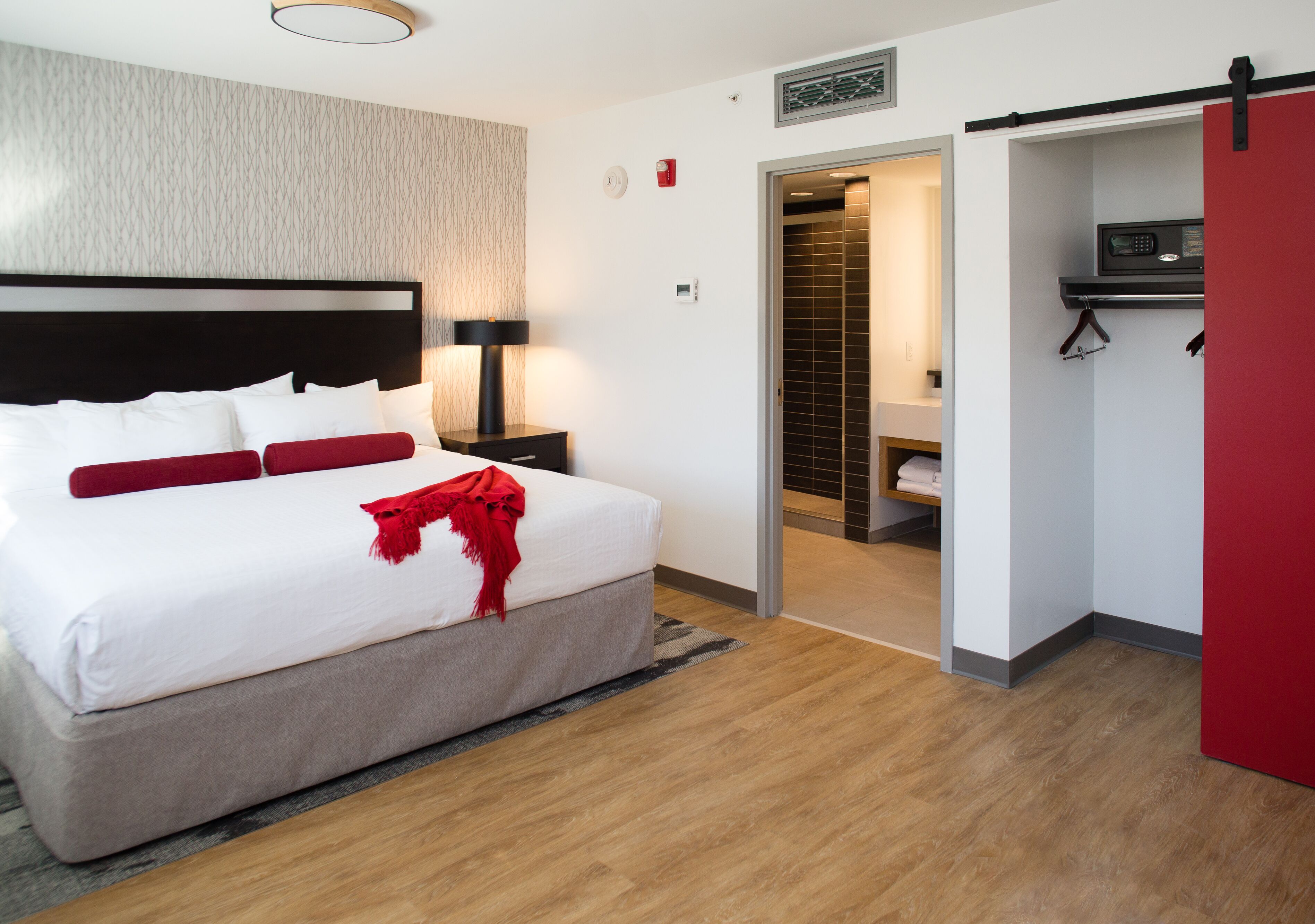 A modern hotel room with a large bed, a bedside table with a lamp, a red door, and a view into a bathroom.
