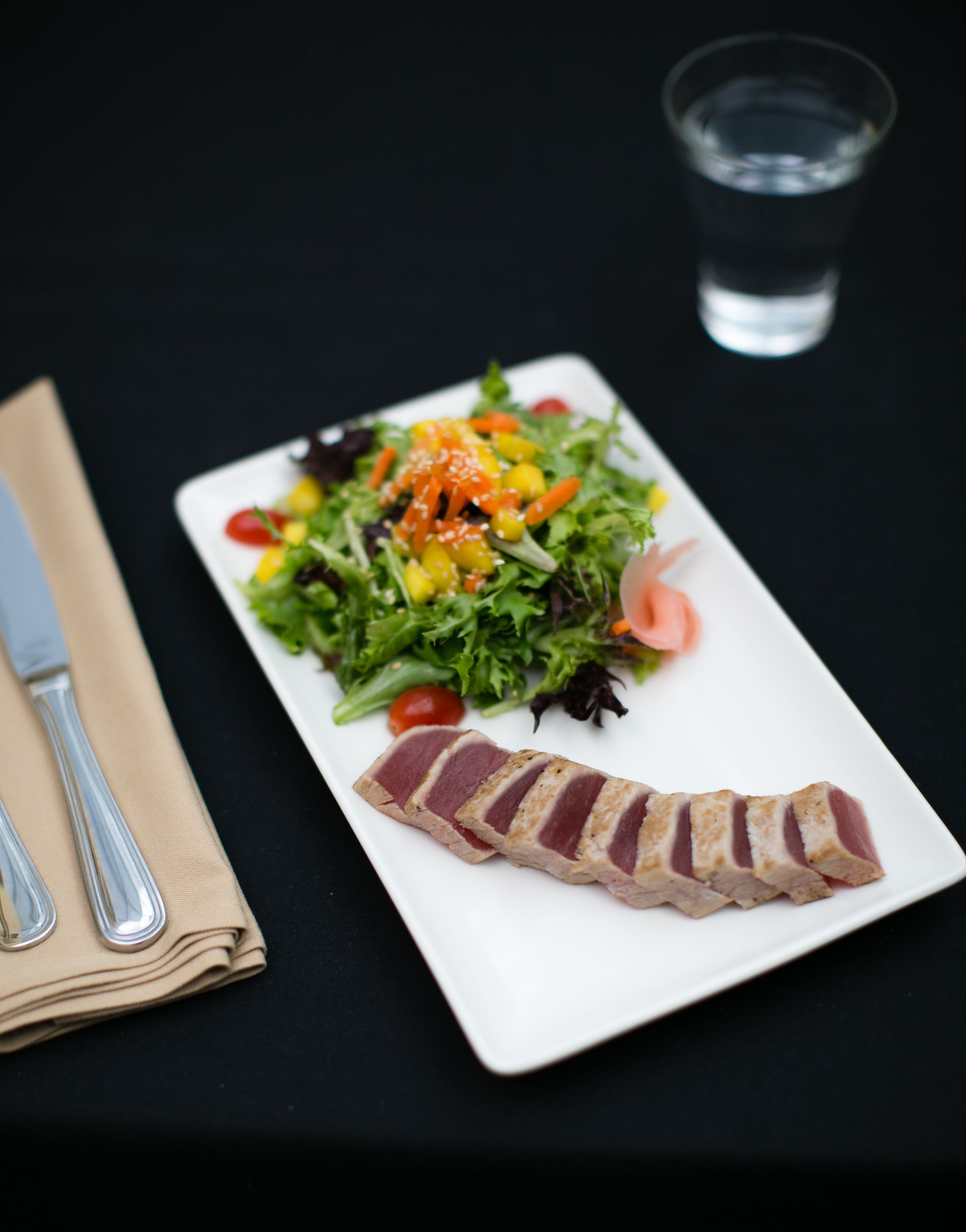 A white rectangular plate with a salad and sliced meat on a black table.