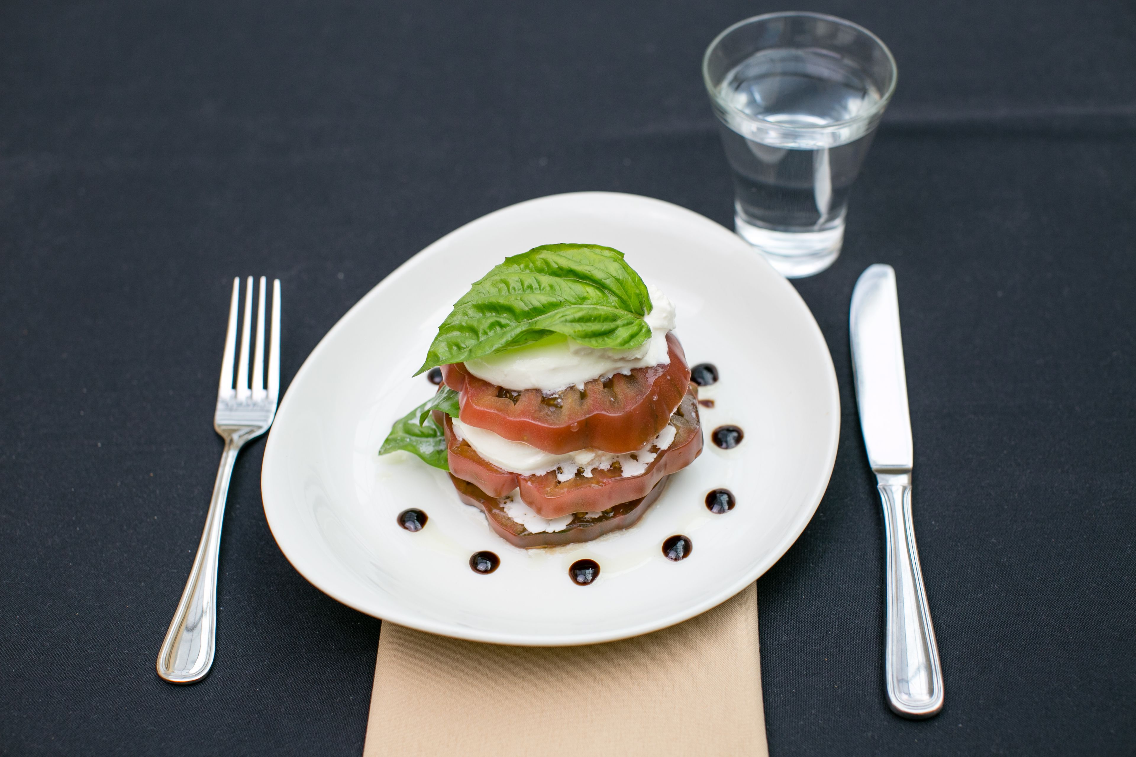 A plate with a layered food item, a fork, a knife, and a glass of water on a dark tablecloth.