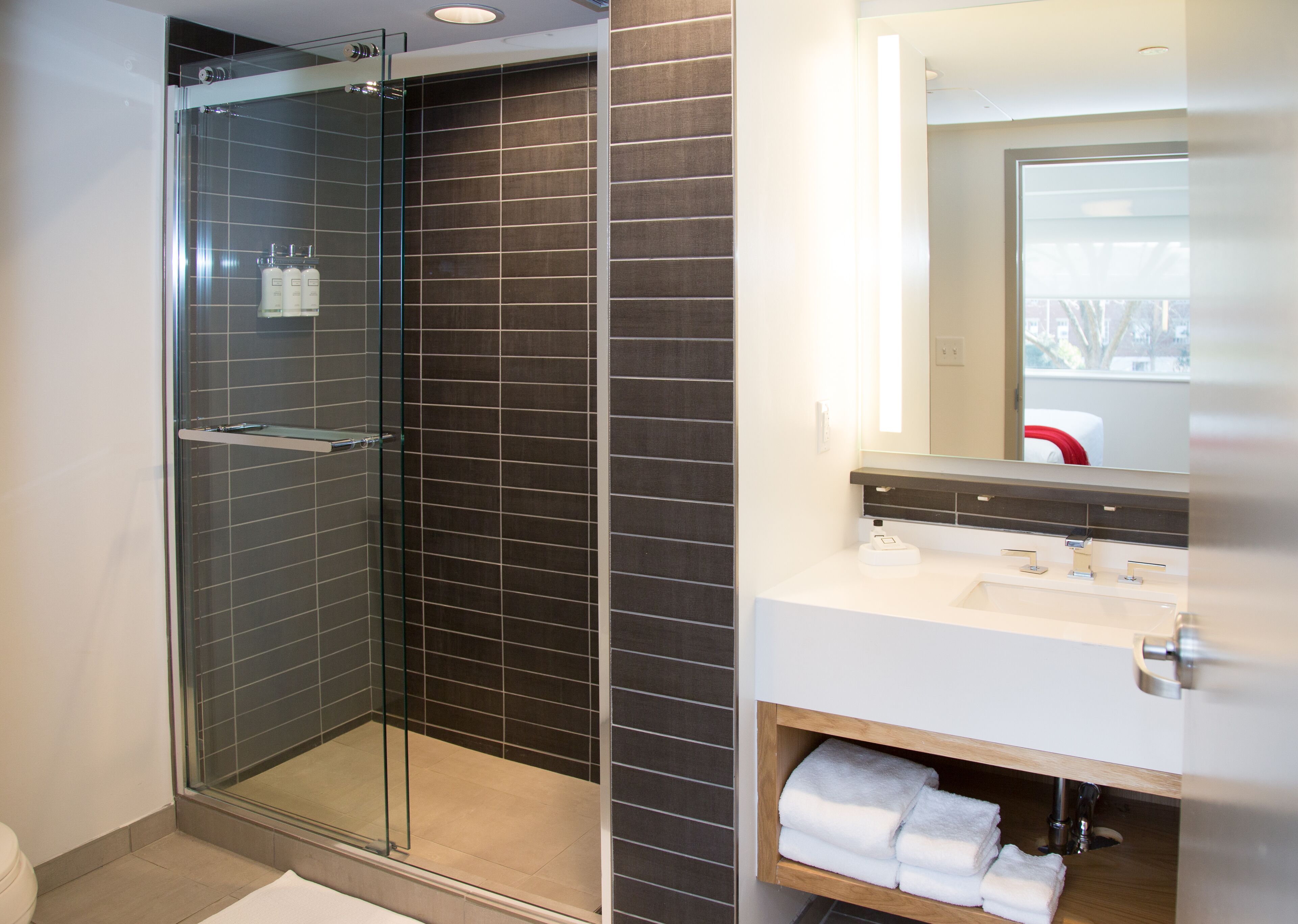 A modern bathroom with a glass-enclosed shower, dark tiled walls, a white sink, and a mirror above the sink.