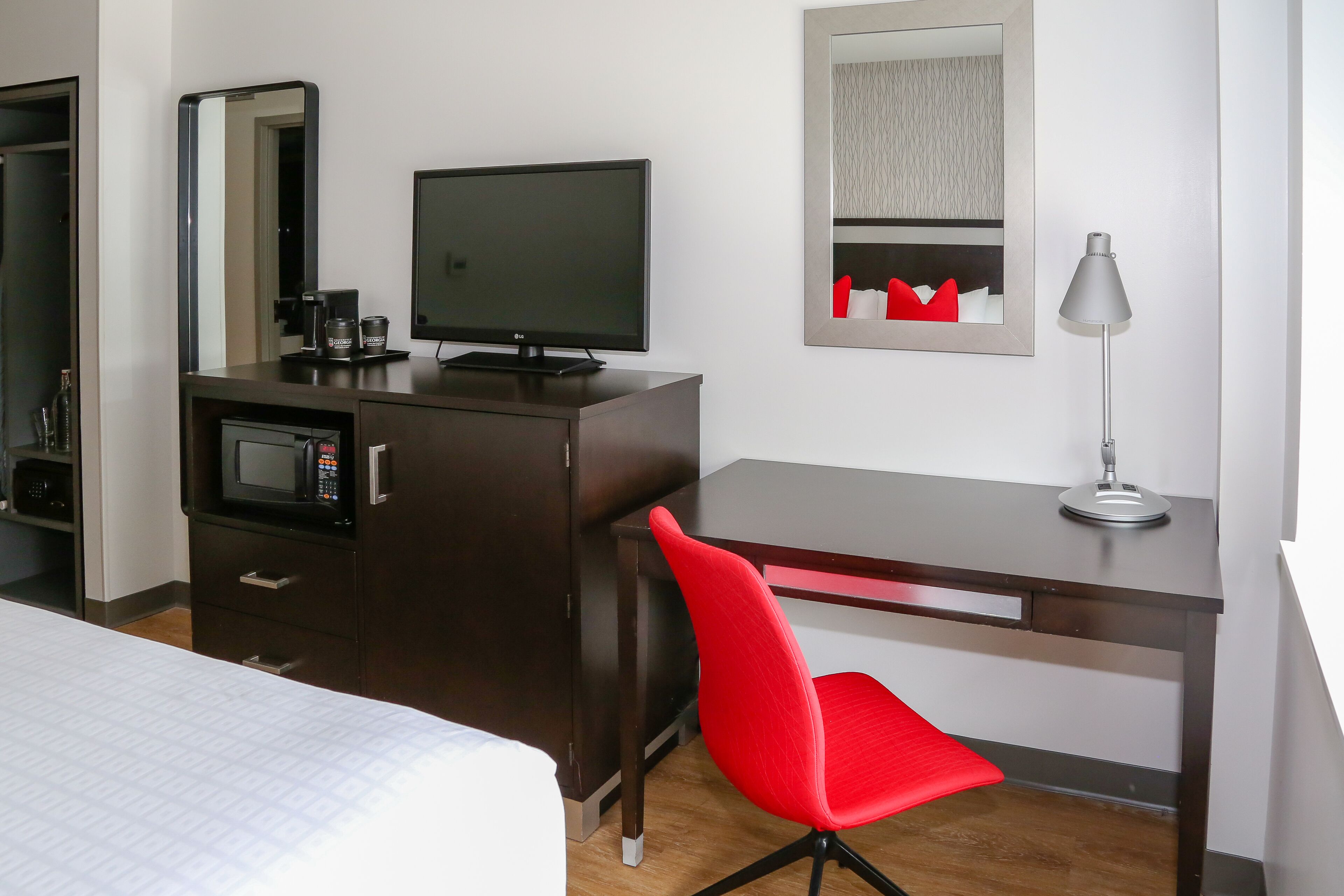 Une chambre d’hôtel avec un lit, un bureau avec un ordinateur, une chaise rouge, une télévision, un four à micro-ondes et une lampe.